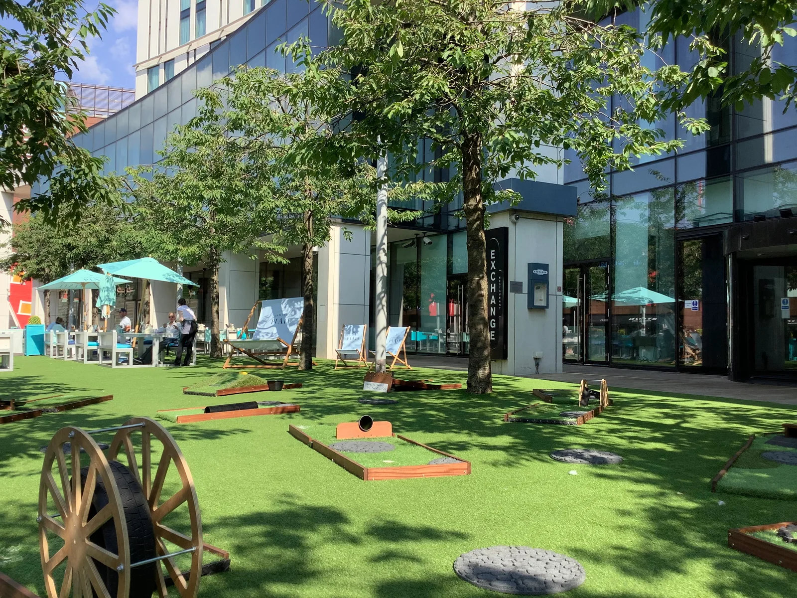 Mini golf holes and wooden obstacles on a green outside a modern glass-fronted venue