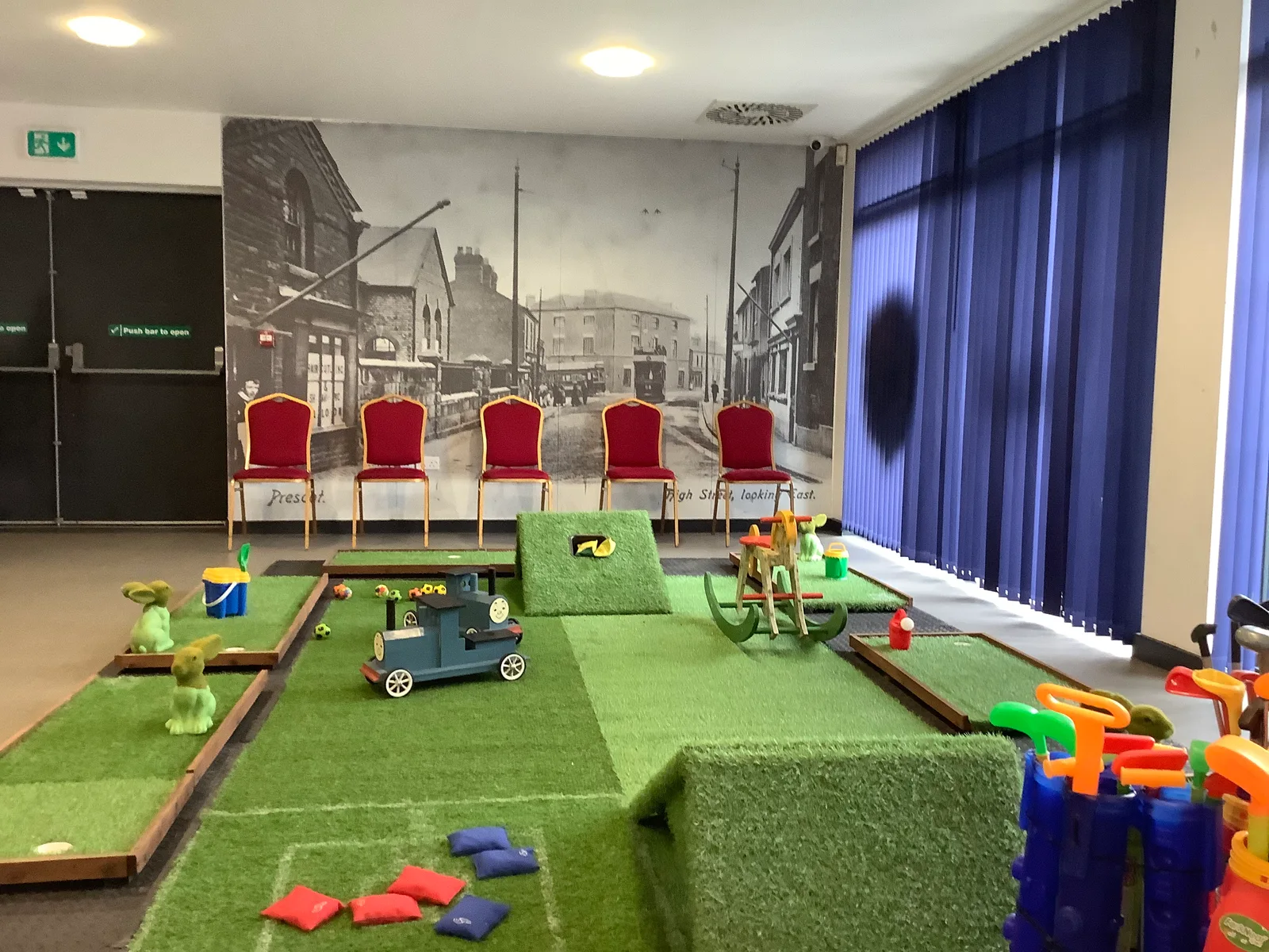 Indoor mini golf course with colourful obstacles, putters and green lanes in a room with blue blinds and red chairs