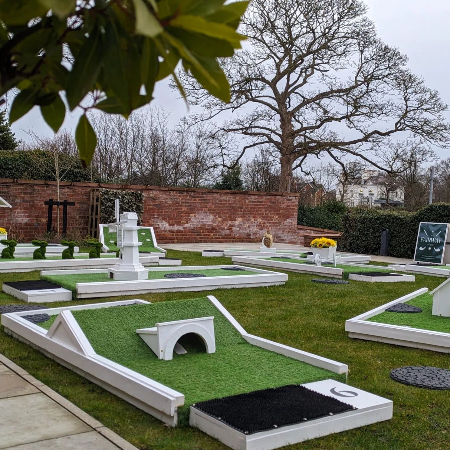 White mini golf holes with a bridge, windmill and topiary obstacles on a garden lawn