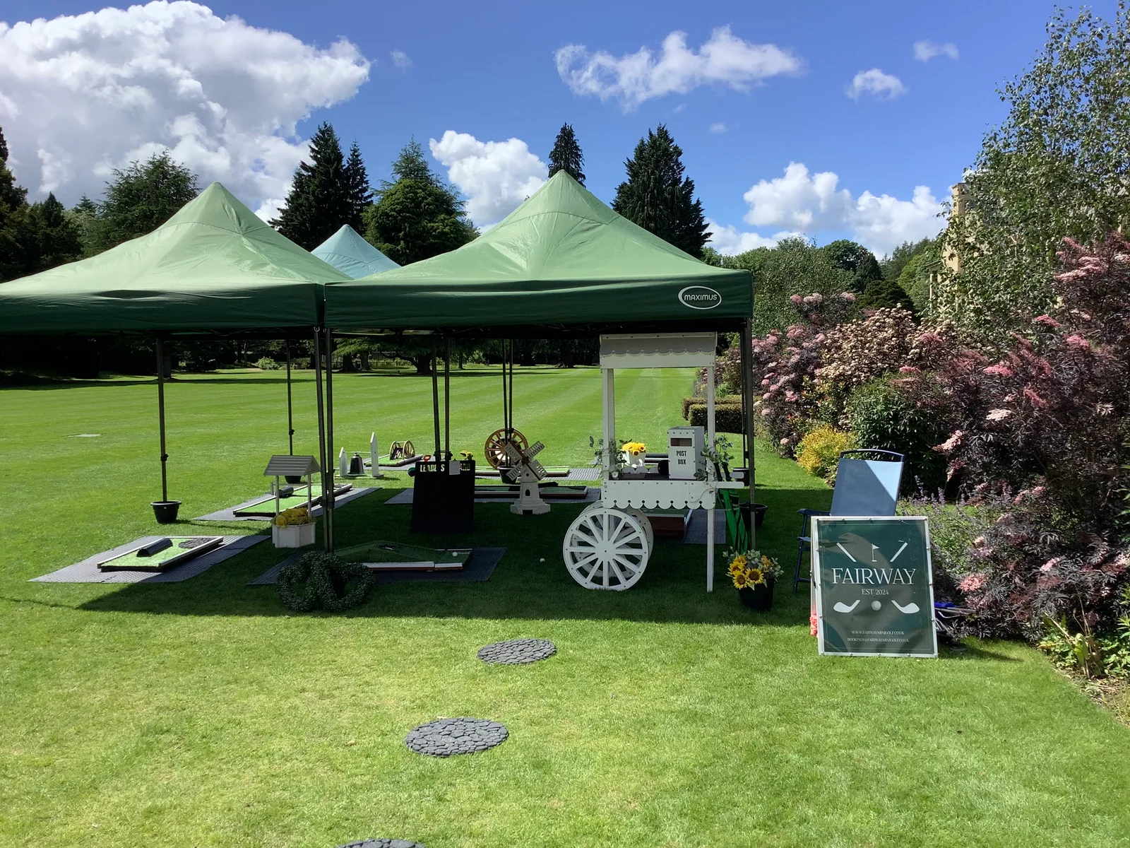 Fairway mini golf beneath green gazebos on a garden lawn, with a white cart and flowering shrubs